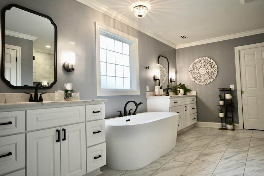 Remodeled bathroom with white cabinets, black fixtures, large white soaking tub between two vanities, under large picture window with grids. Light tile flooring. Gray walls.